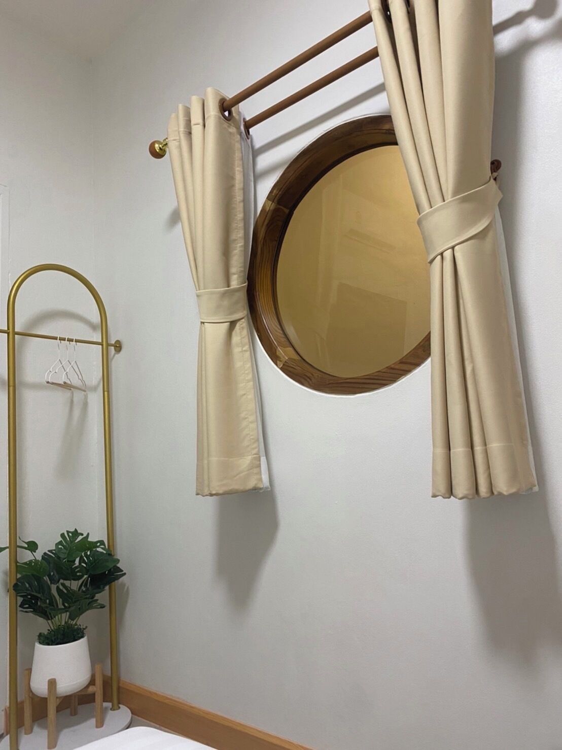 Ein Badezimmer mit einem runden Spiegel, beigen Vorhängen, einem goldenen Metallregal und einer Topfpflanze.