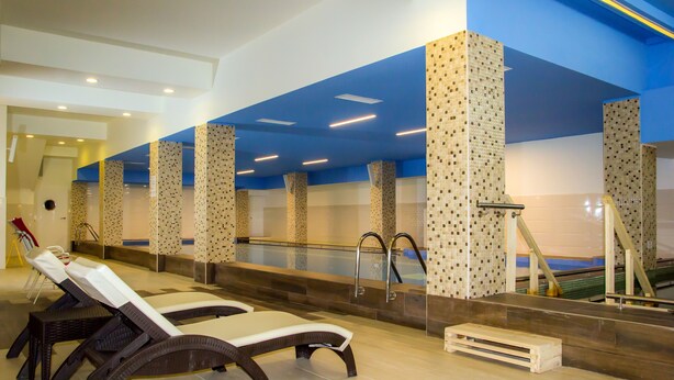 Ein moderner Poolbereich im Innenbereich mit Liegestühlen, einer Glasbarriere und blauer Decke.