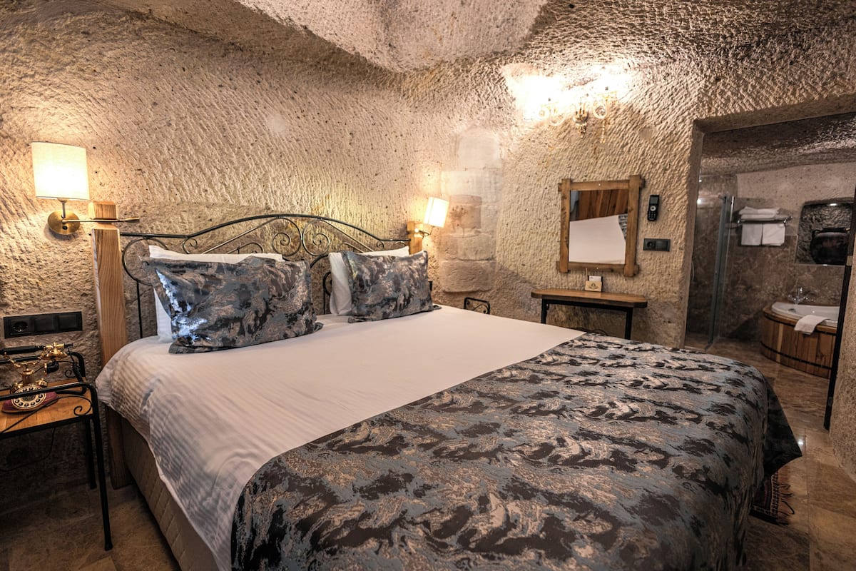 Une chambre avec un mur en pierre, un lit recouvert d’une couvre-lit à motifs, une table de chevet avec une lampe et une tête de lit en bois.