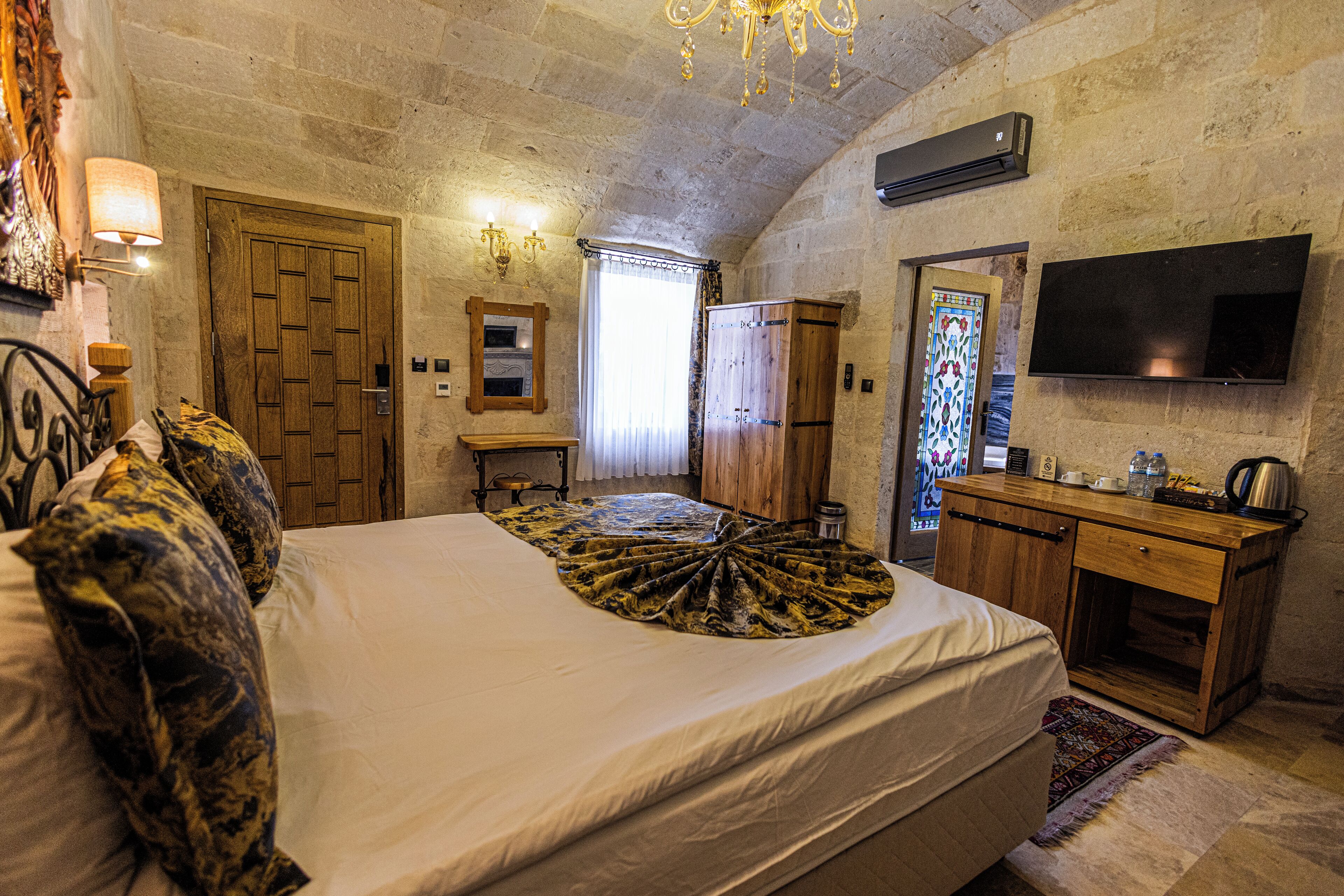 A bedroom with a large bed, a wooden desk, a television, and a stone wall.