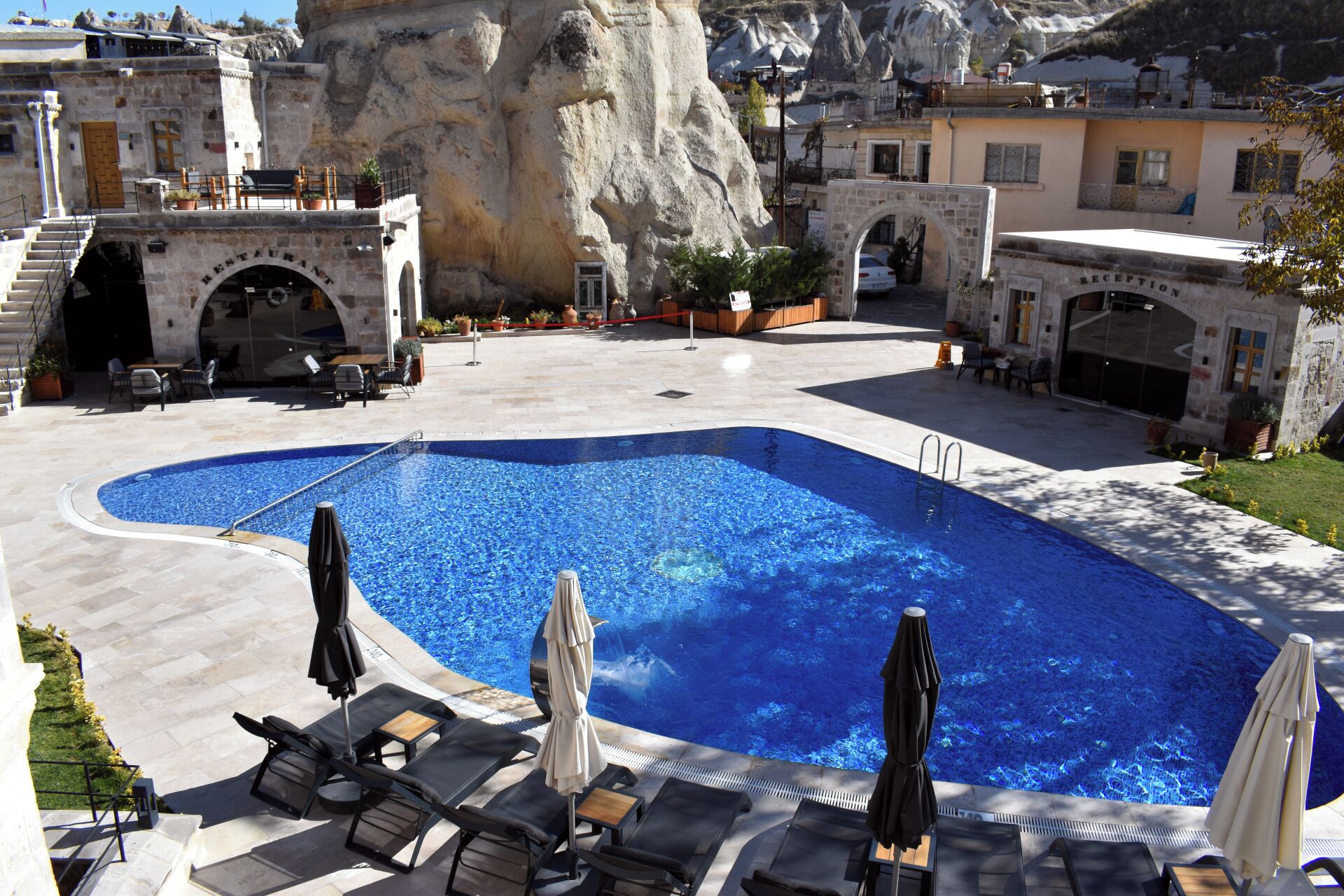 Un espace piscine doté de chaises longues et de parasols, entouré de bâtiments en pierre et avec une chaîne de montagnes en toile de fond.