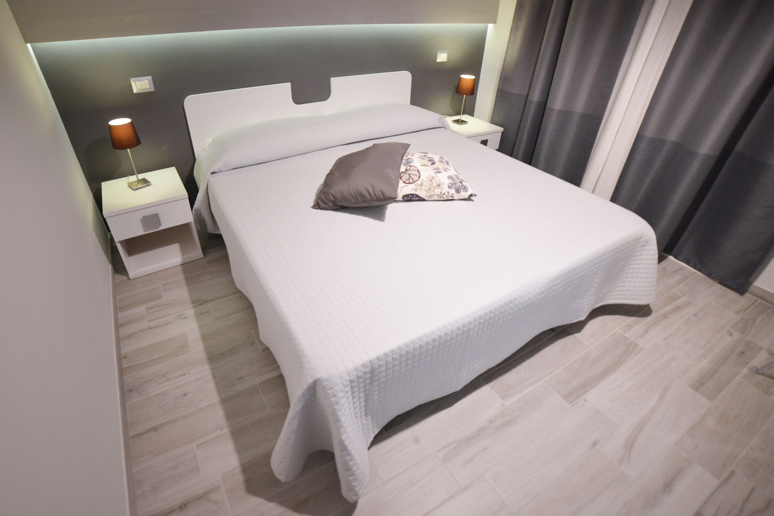 Ein ordentlich bezogenes Bett mit weißem Kopfteil, ein Nachttisch mit Lampe und ein modernes Zimmerdesign.