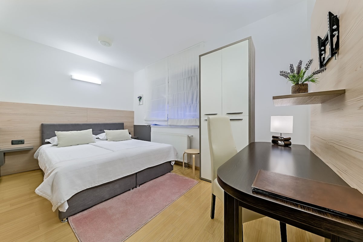 Una habitación de hotel moderna con una cama, mesita de noche, silla y un escritorio pequeño con lámpara.