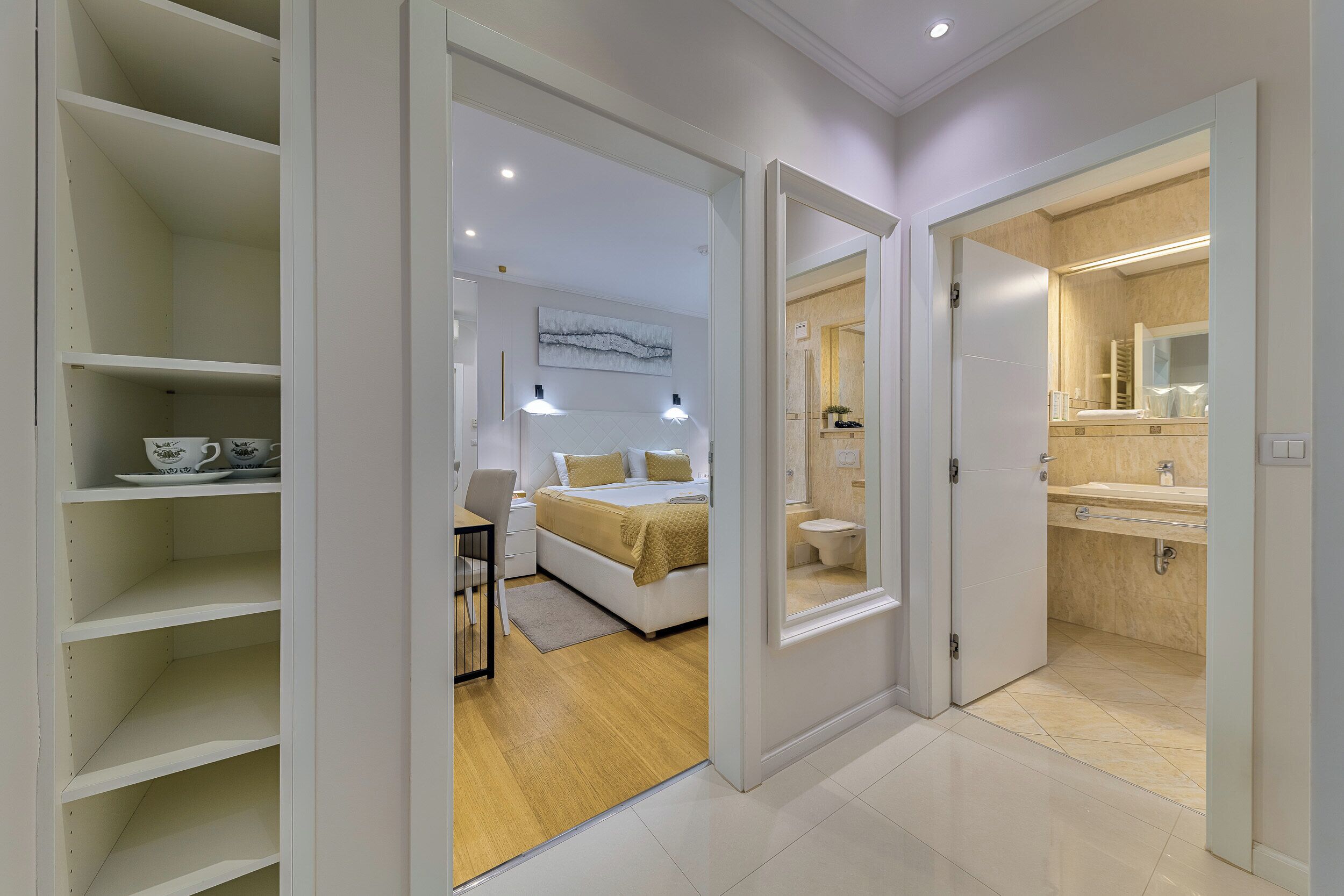 A modern bedroom with a bed, bedside table, and a desk. There is a bathroom with a sink and toilet visible through an open door.