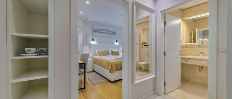 A modern bedroom with a bed, bedside table, and a desk. There is a bathroom with a sink and toilet visible through an open door.