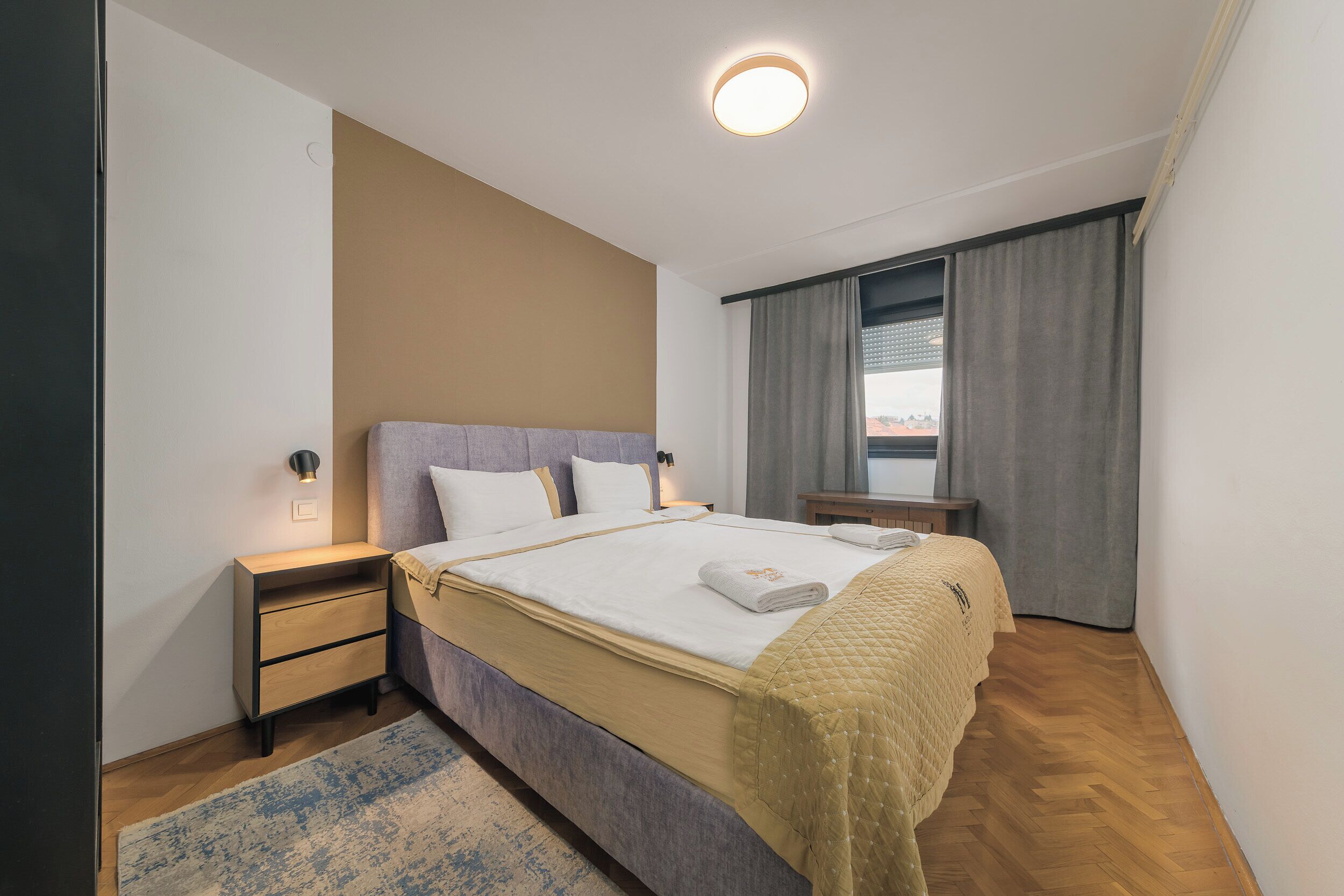 Een moderne hotelkamer met een opgemaakt bed, een nachtkastje, een klein bureau en een raam met gordijnen.