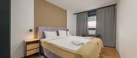 Een moderne hotelkamer met een opgemaakt bed, een nachtkastje, een klein bureau en een raam met gordijnen.