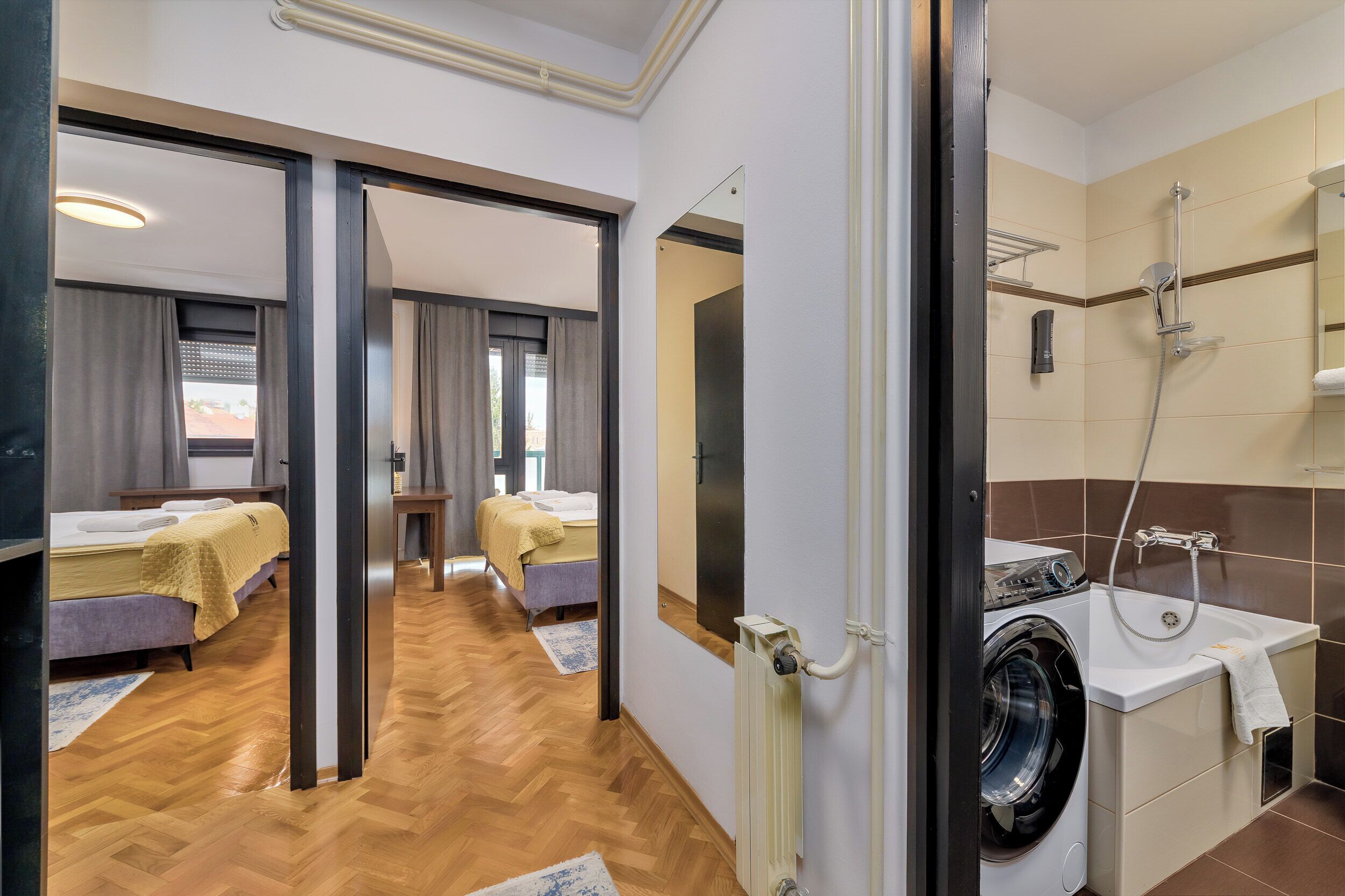 A hotel room with a wooden floor, a bed, a desk, and a bathroom with a washing machine and shower.
