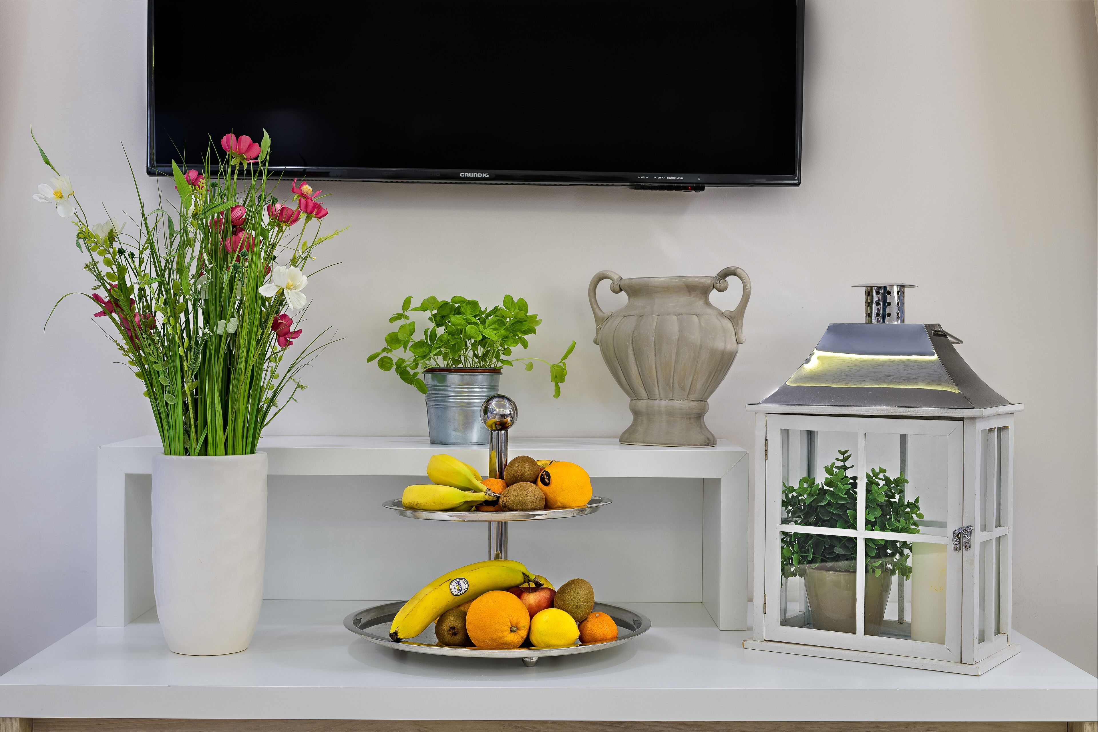 A white table with a fruit platter, a vase of flowers, a metal pot, and a lantern.