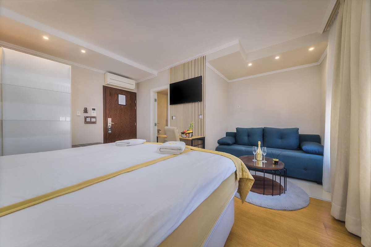 Habitación de hotel con una cama grande, un sofá, una mesita con una botella y vasos, y un televisor montado en la pared.