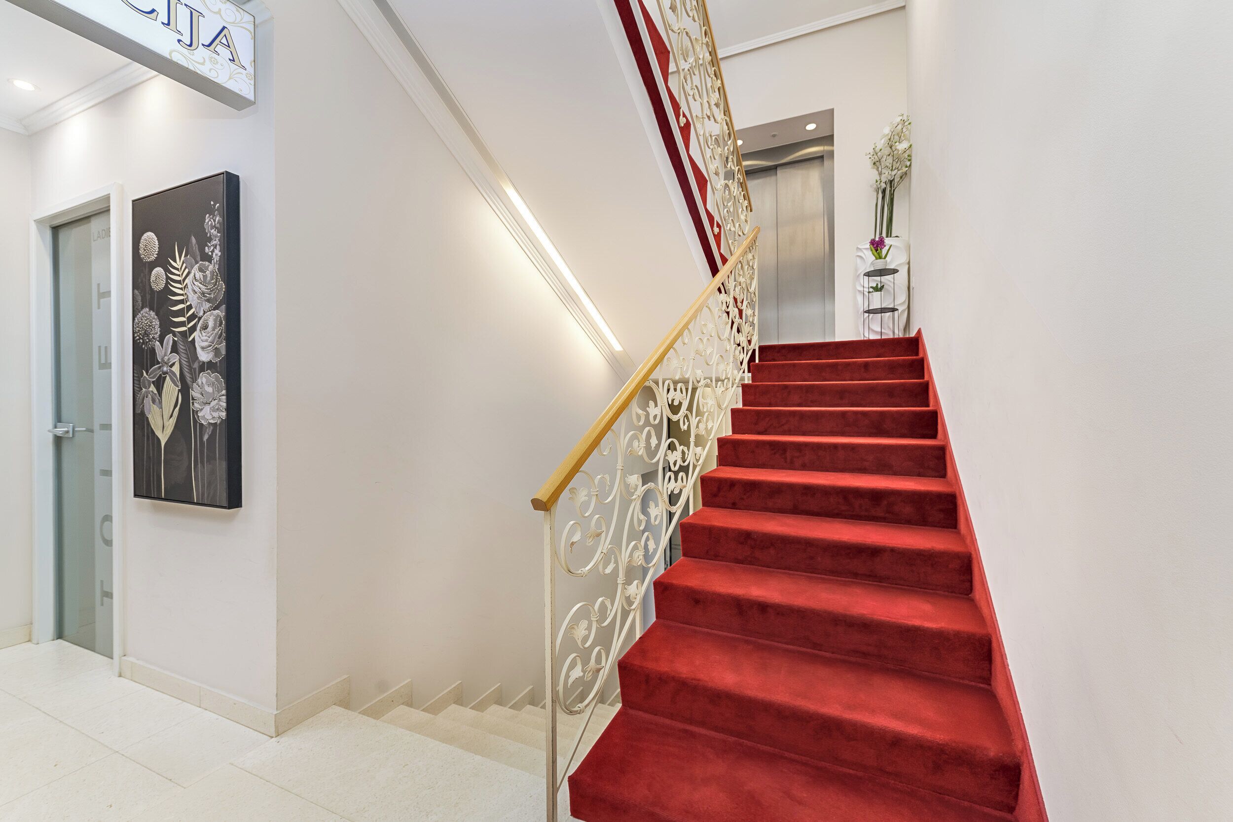 A staircase with red carpet and ornate metal railings, leading to an elevator.