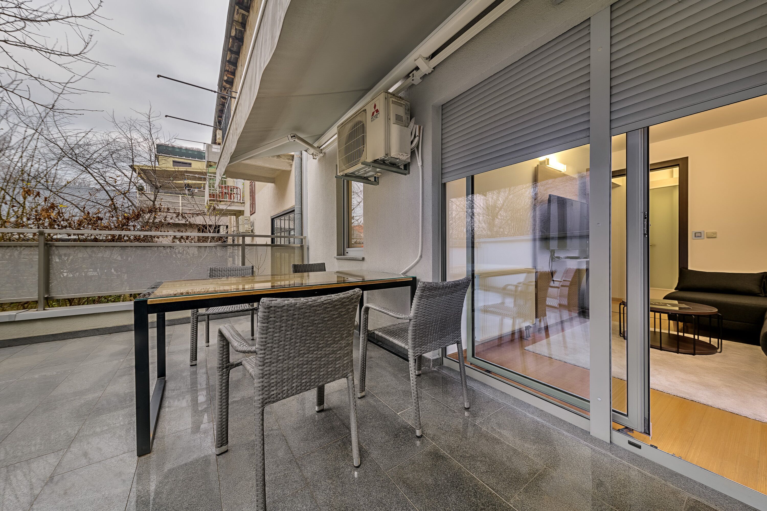 Un balcón al aire libre con una mesa y sillas, una puerta de cristal que da a una sala de estar y vista a un edificio al otro lado de una cerca.