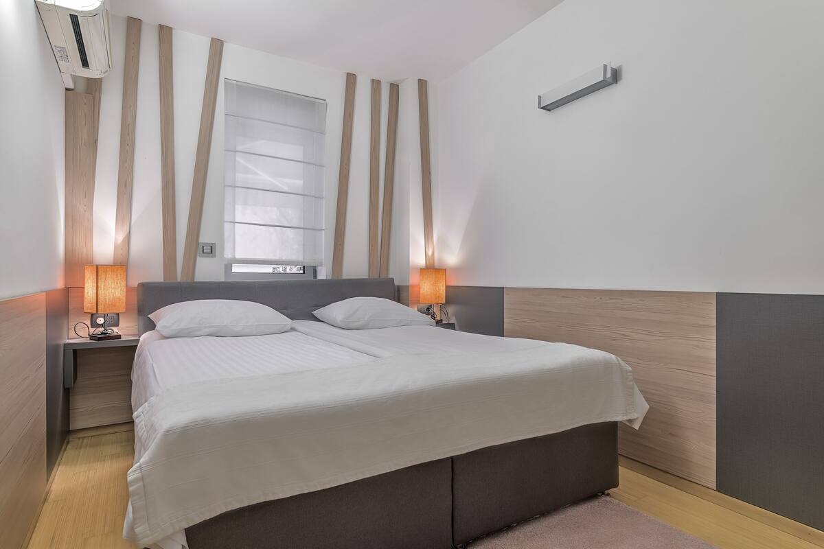 Un dormitorio moderno con una cama grande, cabecera de madera, dos mesitas de noche con lámparas y una ventana con cortinas.