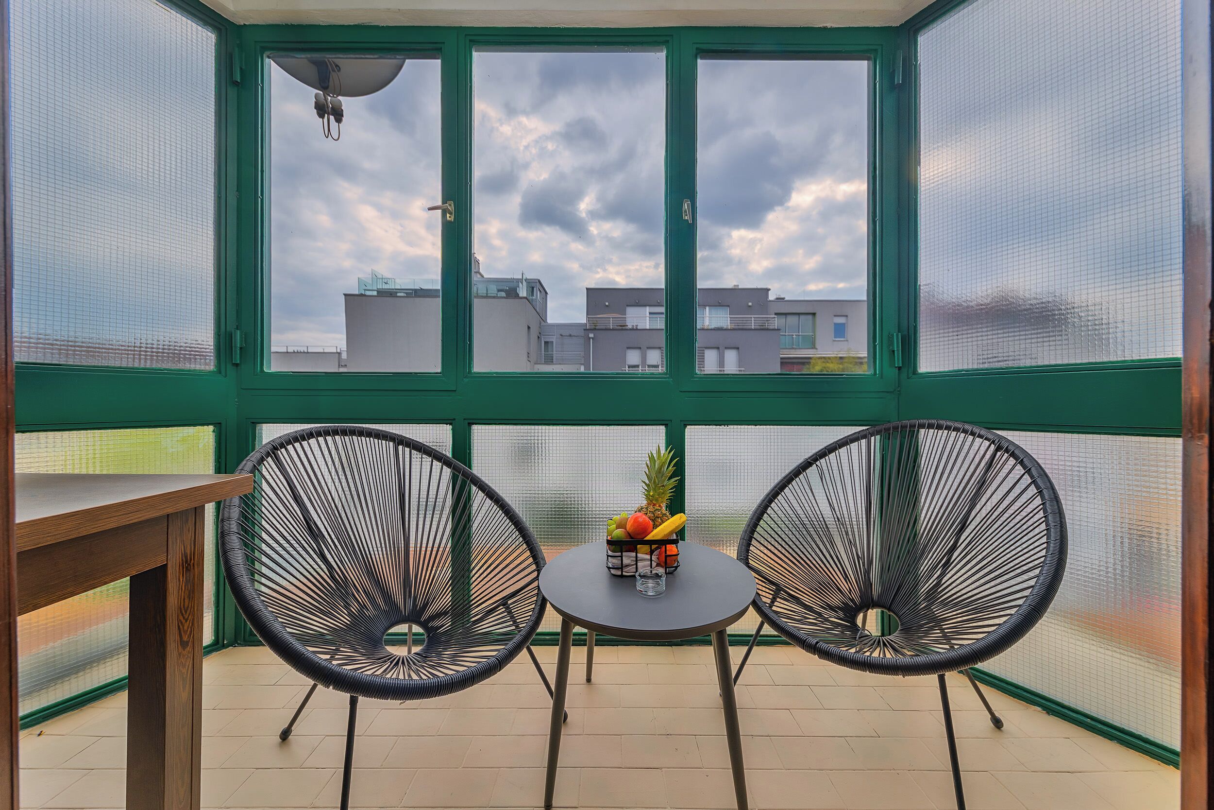 Un balcon avec deux chaises en fil de fer noir, une petite table ronde sur laquelle se trouve un panier de fruits, et une vue sur les bâtiments, visible à travers une fenêtre à cadre vert.