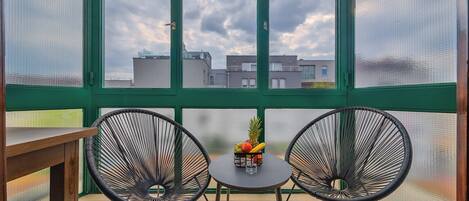 Un balcon avec deux chaises en fil de fer noir, une petite table ronde sur laquelle se trouve un panier de fruits, et une vue sur les bâtiments, visible à travers une fenêtre à cadre vert.