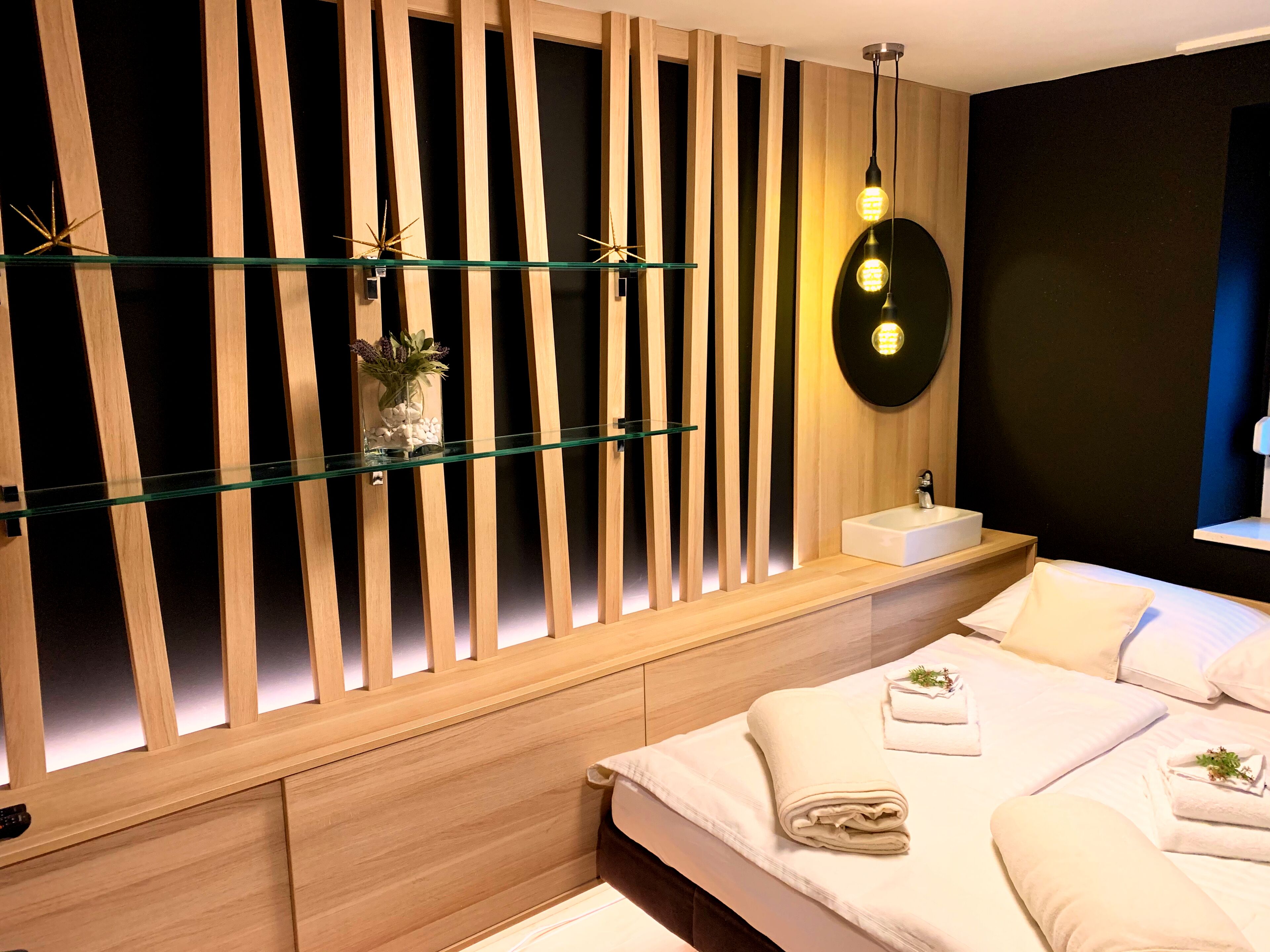 A modern bedroom with a wooden slat wall, a glass shelf with decorative items, a bedside table with a sink, and neatly made beds with towels.