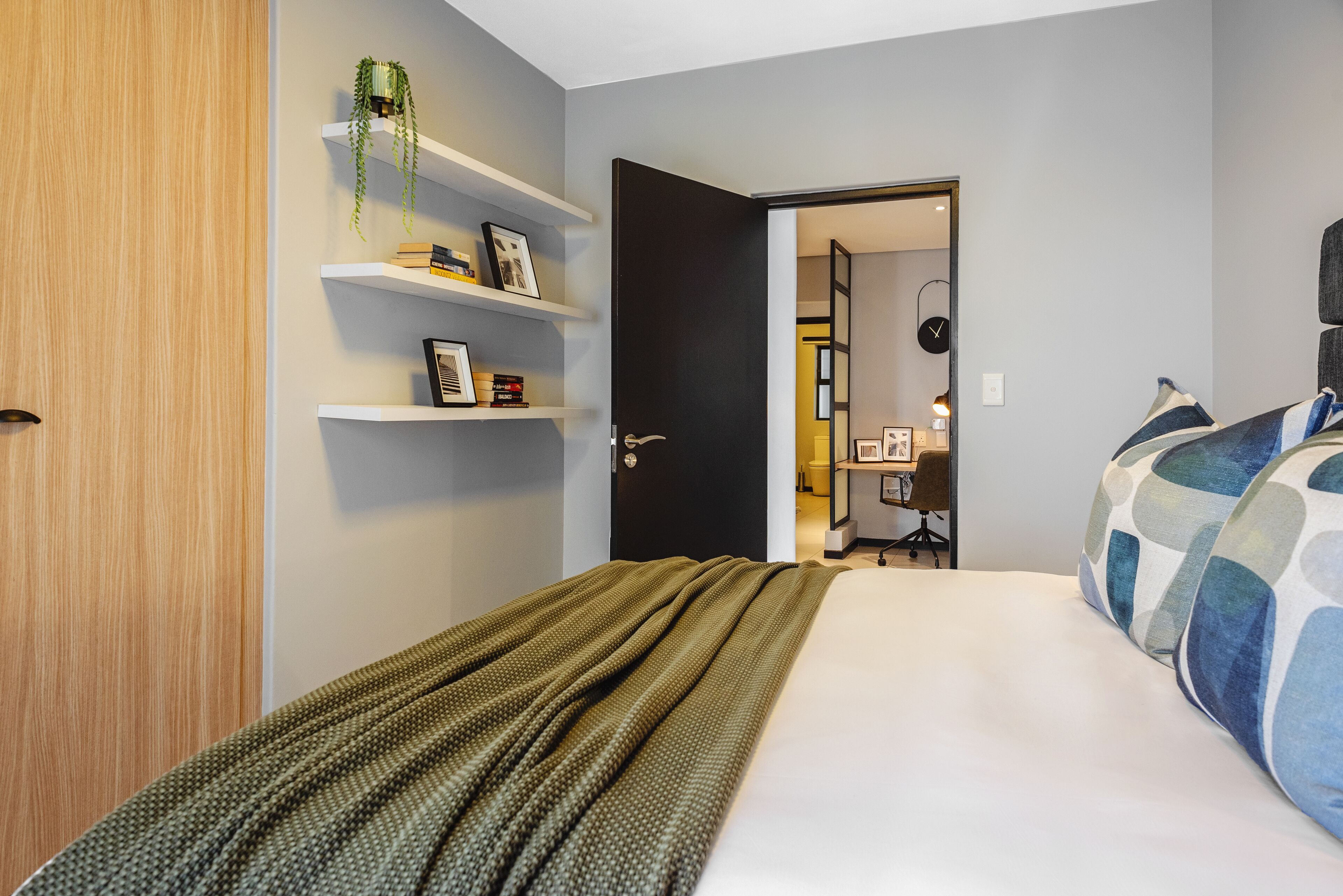 Ein Schlafzimmer mit einem Bett, einer Holztür, einem Regal mit Büchern und einer Pflanze sowie einem Schreibtisch und einem Stuhl im Hintergrund.