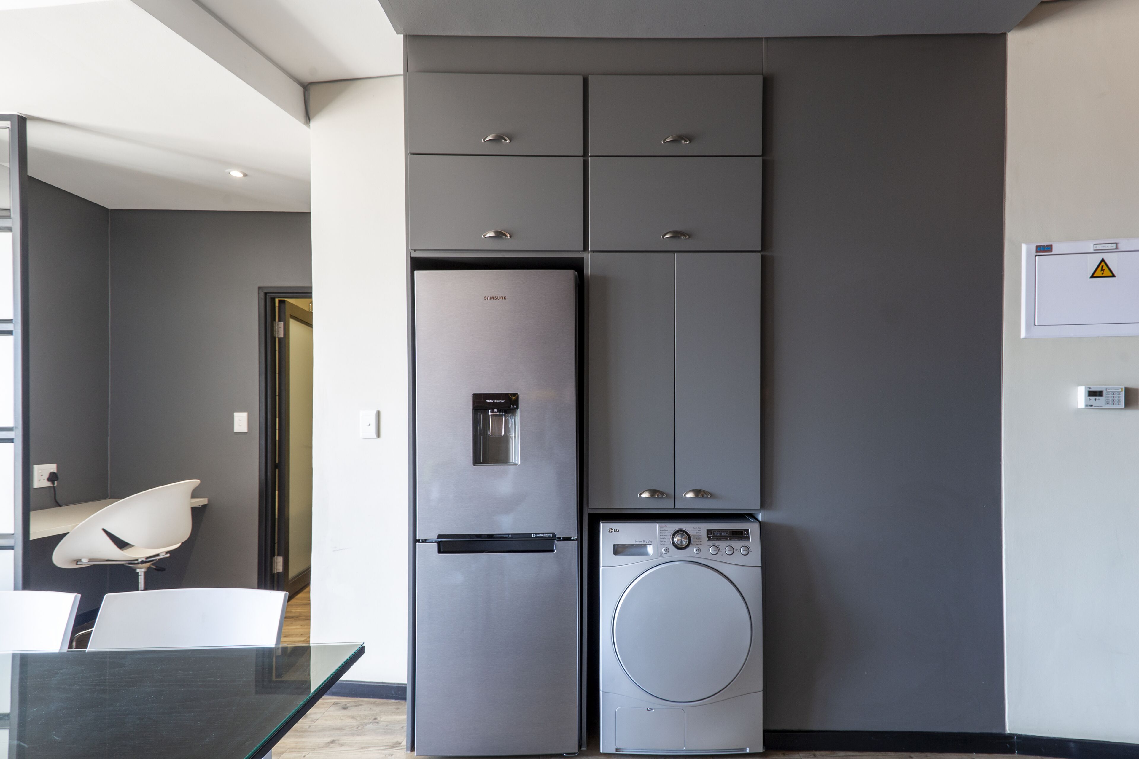 A compact kitchen with a stainless steel refrigerator, a built-in washing machine, and a glass dining table with chairs.