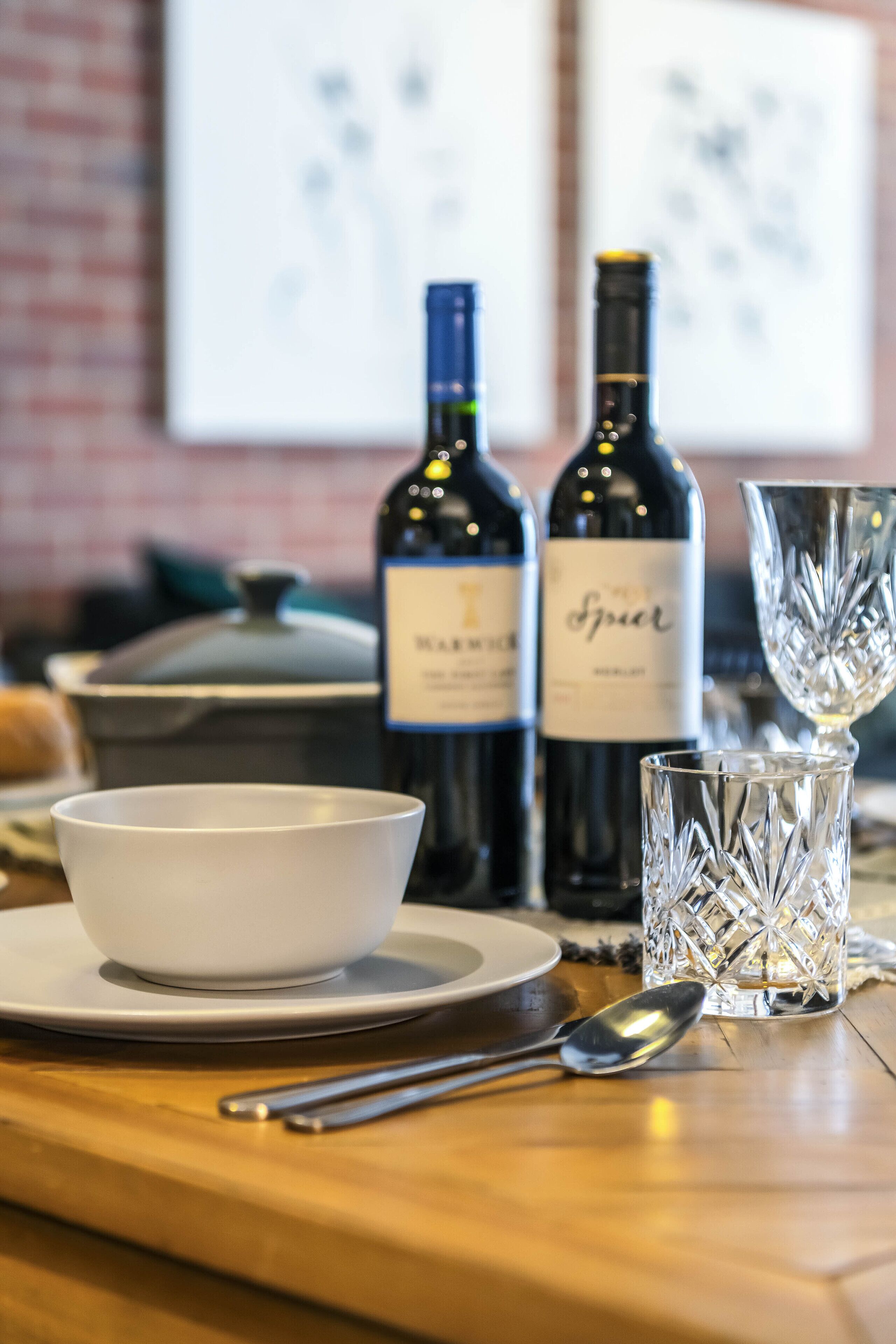 Una mesa de comedor con un tazón blanco, una botella de vino, una copa de vino y una cuchara.