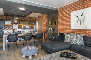 Una cocina moderna con comedor, una sala de estar con sofá y reposapiés, y una pared de ladrillo con obras de arte.