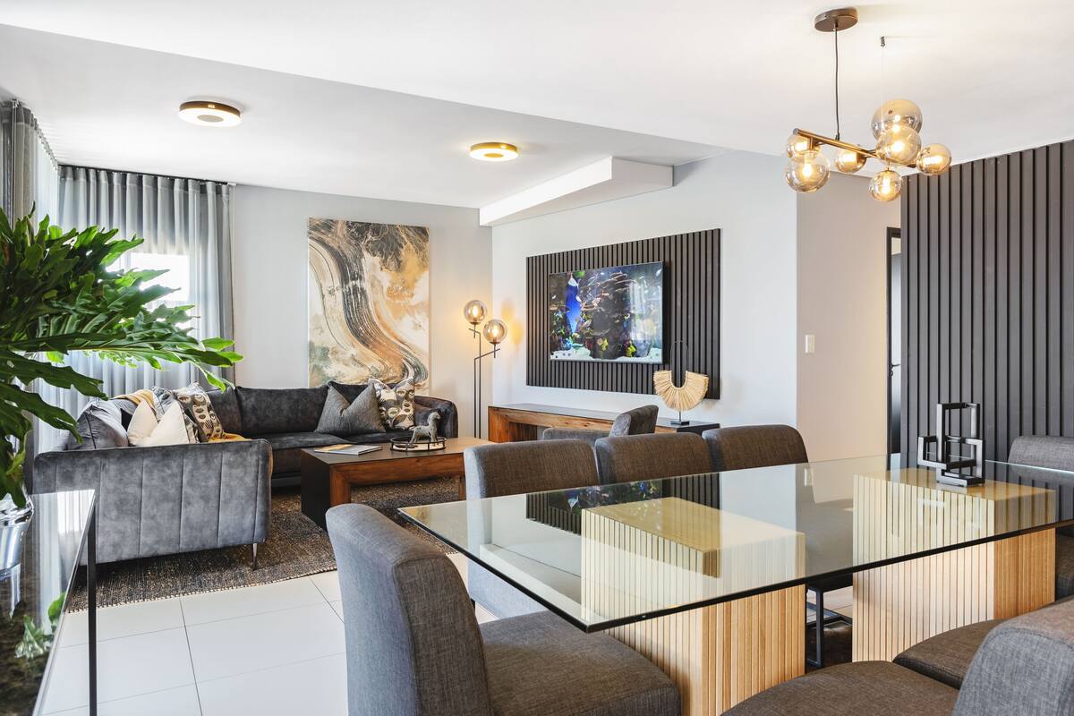 Een moderne woonkamer met een glazen eettafel, grijze banken en een groot abstract schilderij.