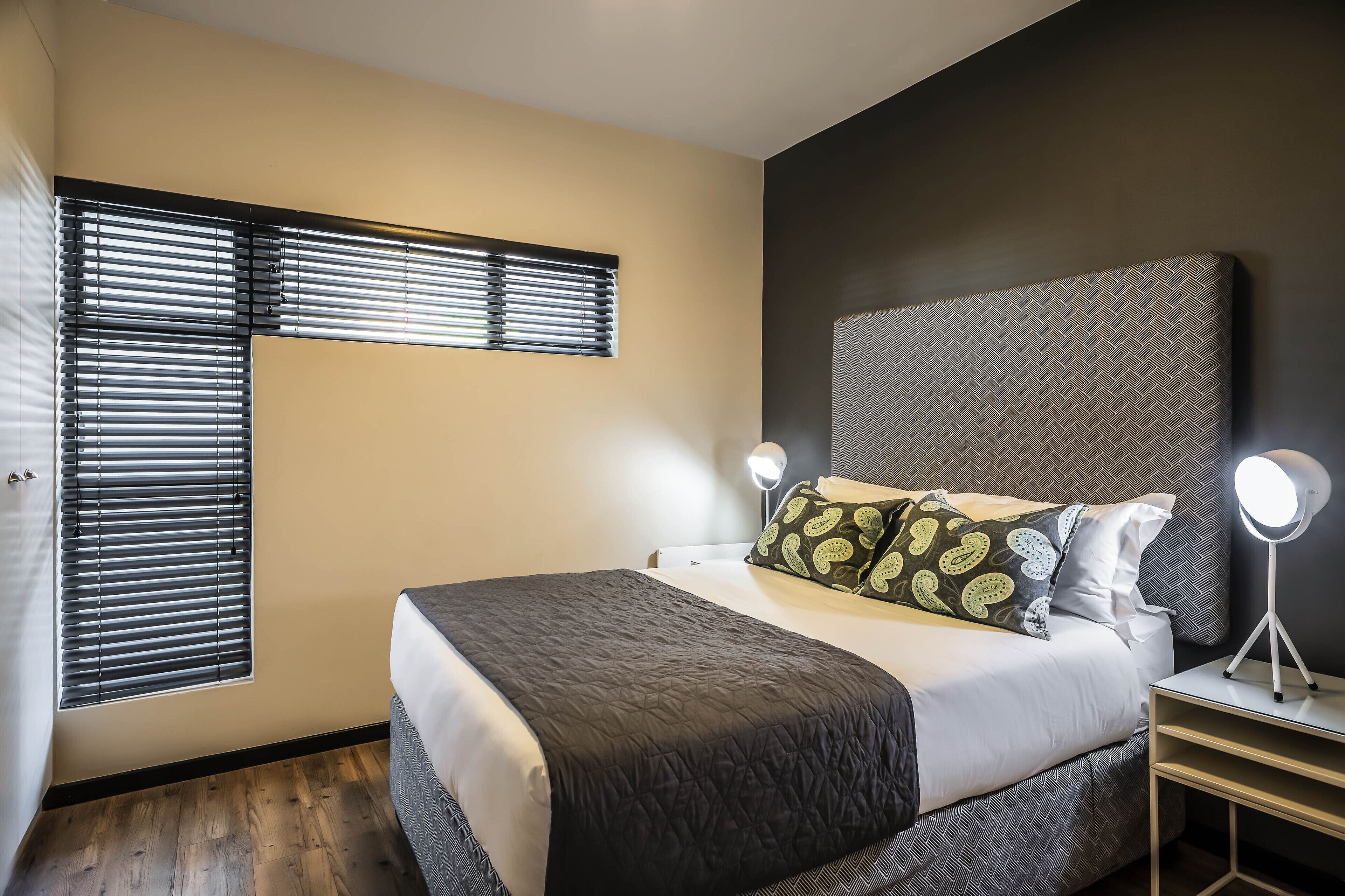 A bedroom with a bed, bedside lamps, a window with blinds, and a headboard.
