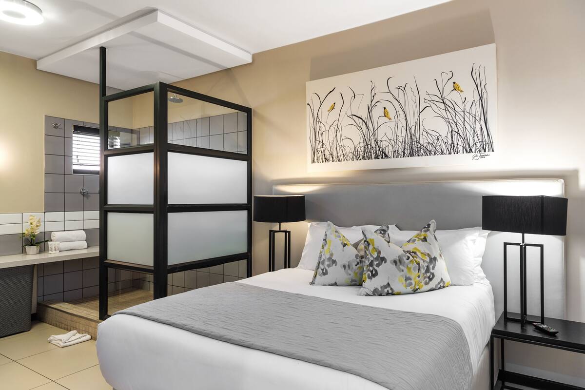 Een moderne hotelkamer met een bed, nachtkastjes, een badkamer met douche en een groot abstract schilderij aan de muur.