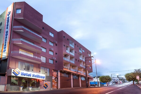 Um hotel com vários andares e varandas, um letreiro com a inscrição 'Hotel Solimano', e uma vista da rua com um ônibus azul e árvores.