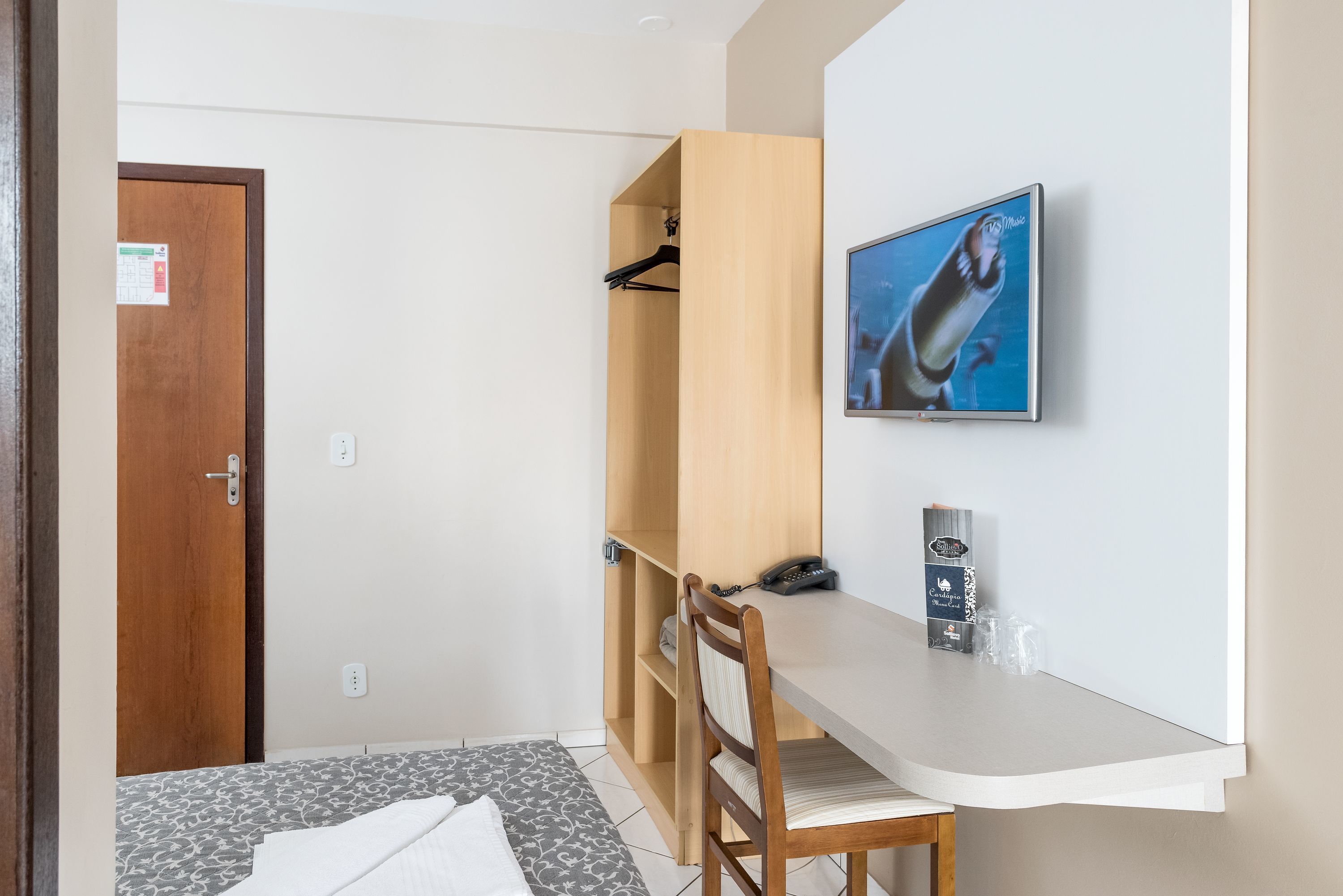 A hotel room with a bed, a desk with a chair, a wooden wardrobe, a wall-mounted TV, and a door.