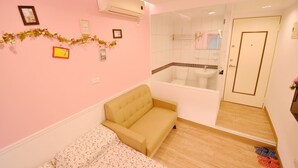 Une chambre à coucher avec un canapé beige, un lit recouvert d’une couvre-lit à motifs floraux, une salle de bain avec une baignoire et un miroir.