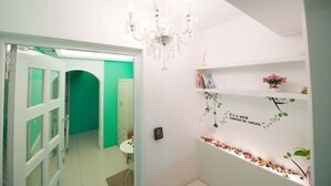 Un couloir avec une porte verte, un mur blanc orné d’une étagère décorative pour plantes et un lustre.