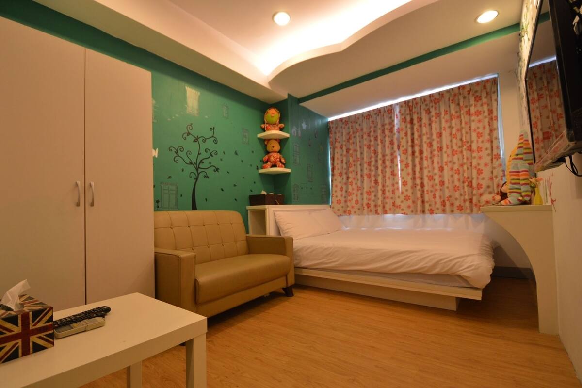 A bedroom with a bed, a sofa, a table with a remote control, and a teddy bear on a shelf.