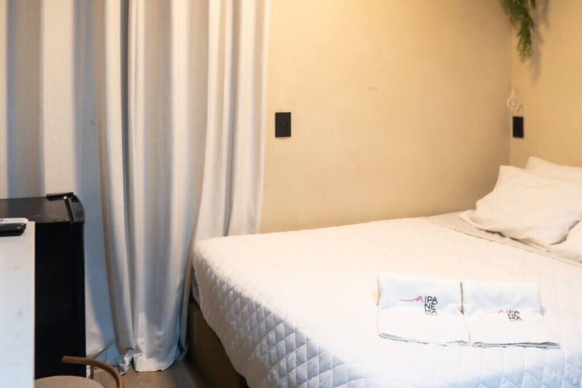 Un dormitorio con una cama, cortinas blancas, una planta pequeña y un cubo de basura blanco.