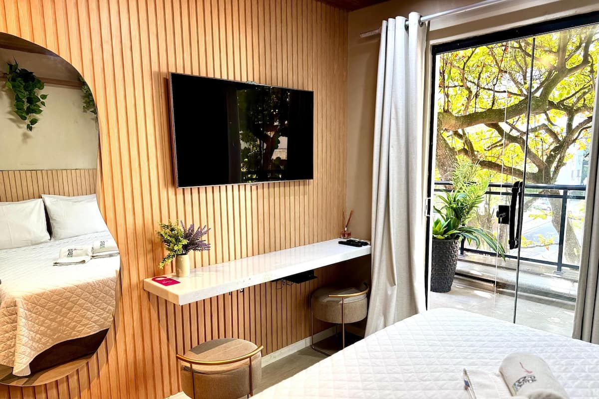 Un hotel moderno con cama, televisor montado en la pared, un escritorio pequeño con silla y ventana con vistas a árboles.