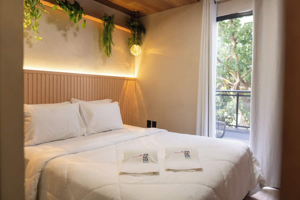Una cama bien tendida con ropa blanca y dos toallas con un logotipo, una planta colgante y una lámpara de pared.