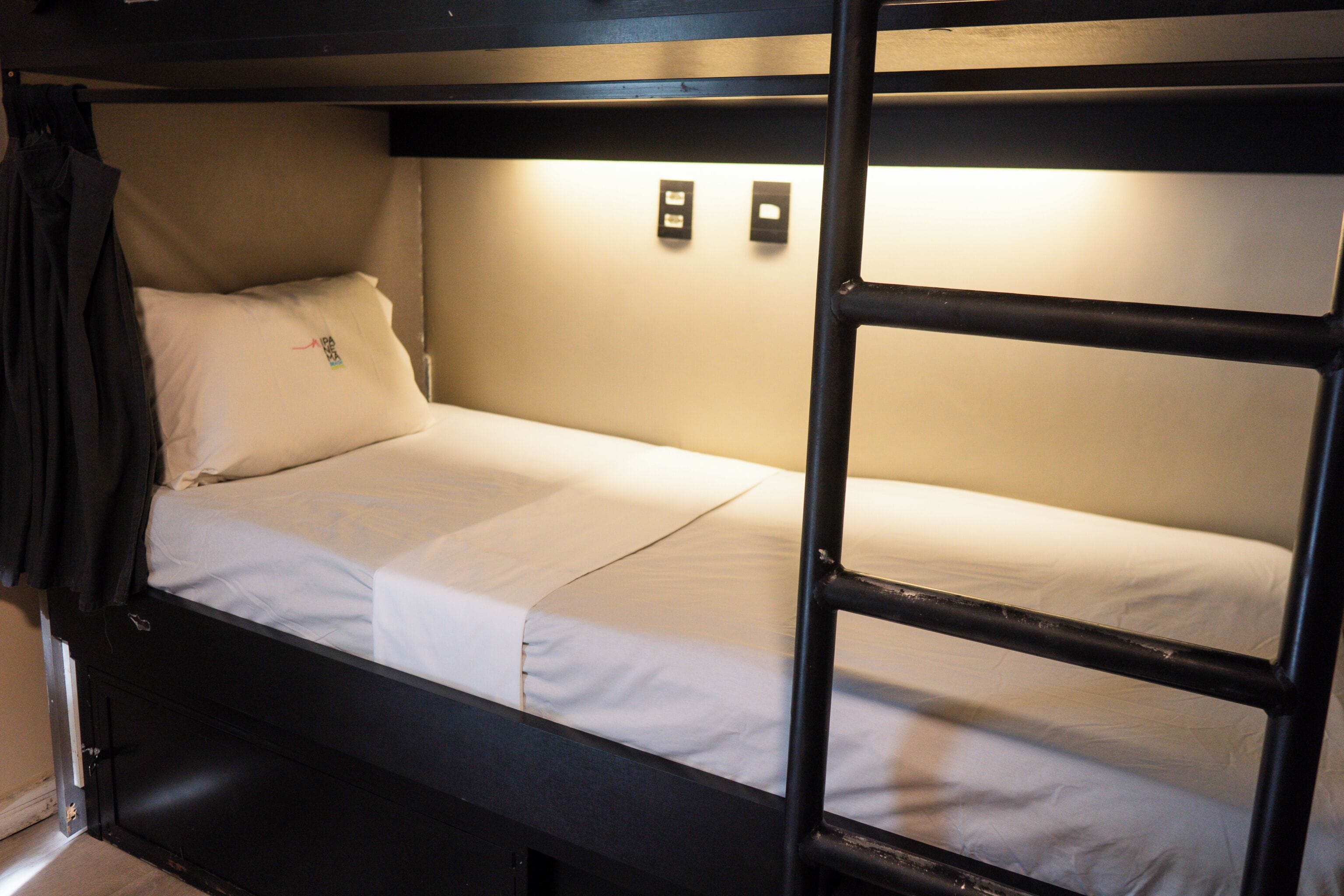 A bunk bed with a white mattress and a black frame. There are two wall-mounted light switches and a black jacket hanging on the left side of the bed.