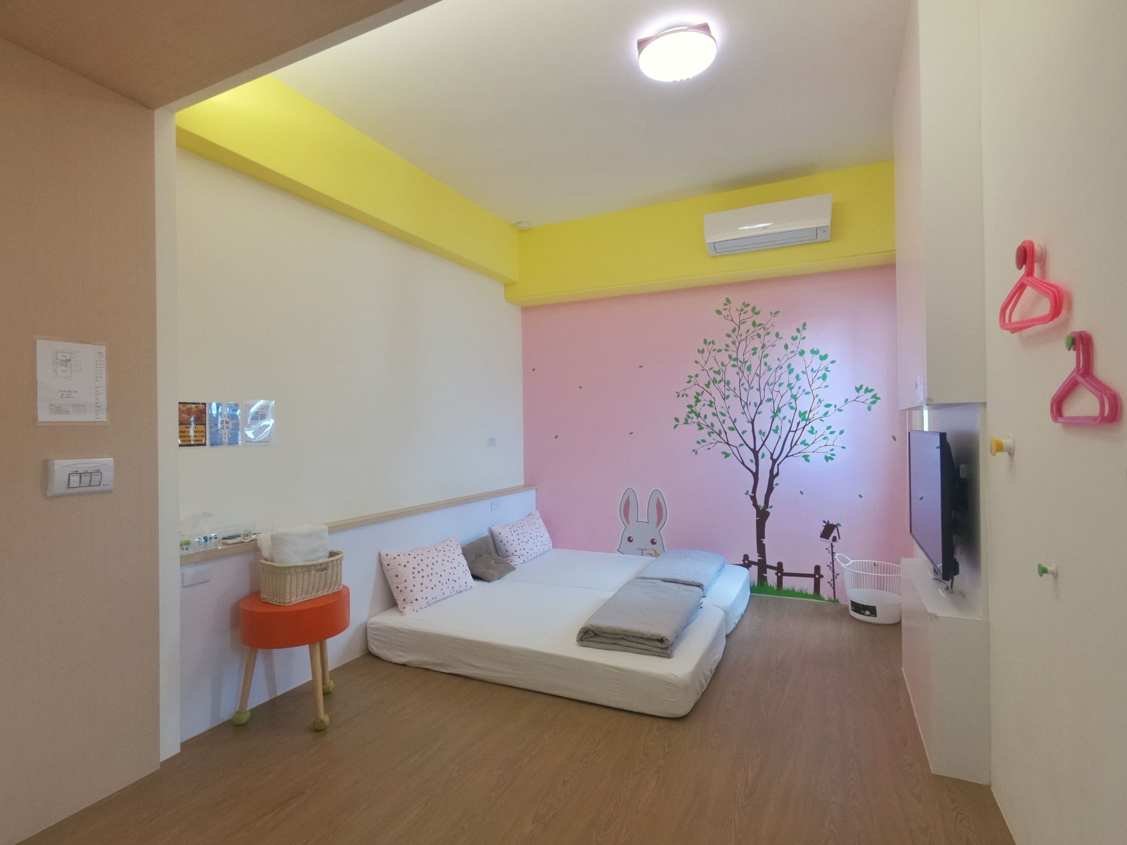 A bedroom with a bed, a wall-mounted air conditioner, a small table with a basket, and a wall decoration of a tree and rabbit.