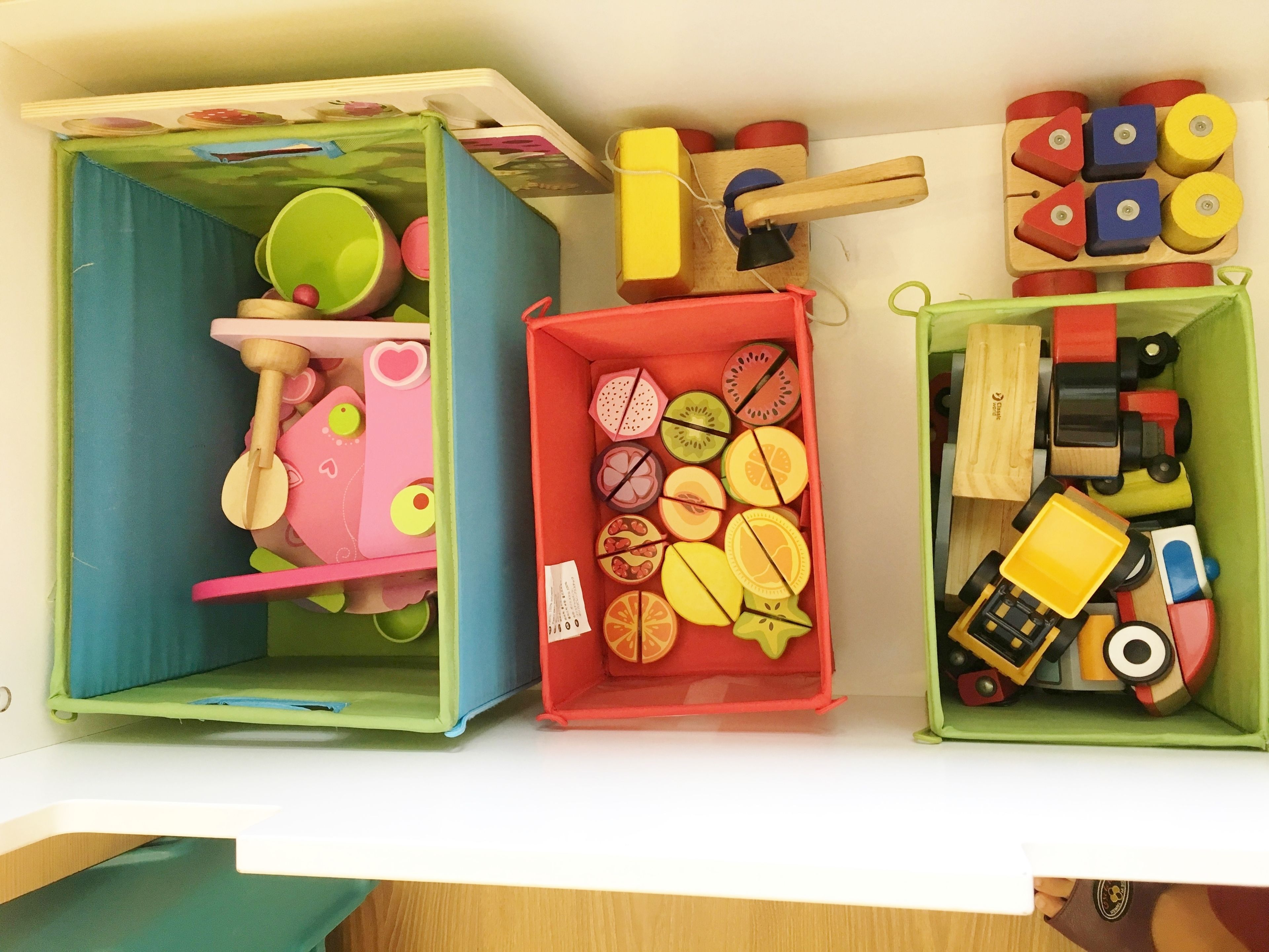 Three storage boxes with various items inside, including wooden toys and fruit-shaped objects.