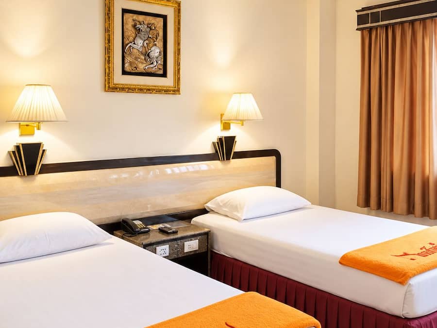 Una habitación de hotel con dos camas, una mesita de noche con teléfono, un cuadro enmarcado en la pared y un ventilador de techo.