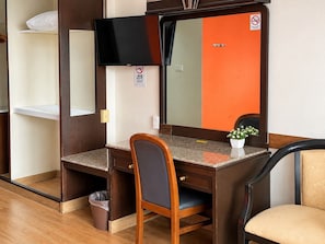 Une chambre d’hôtel avec un bureau en bois, une chaise, une télévision, un miroir et une petite plante en pot.