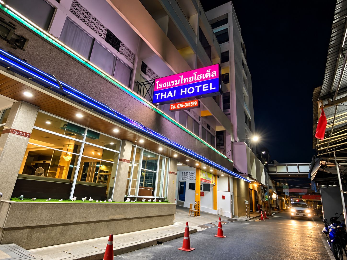 Ein Hotel mit einer Neonreklame, einem sichtbaren Eingang und einem Straßenbild mit Verkehrskegeln.
