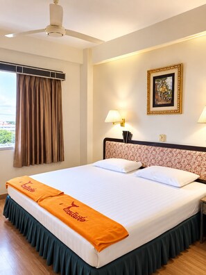 Une chambre d’hôtel avec un lit, un ventilateur de plafond, une fenêtre avec des rideaux, une lampe fixée au mur et un tableau encadré.