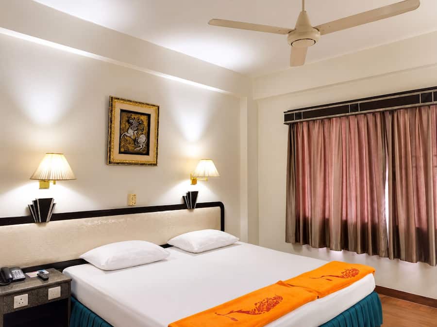A hotel room with a bed, two bedside lamps, a ceiling fan, a framed picture on the wall, and a window with curtains.
