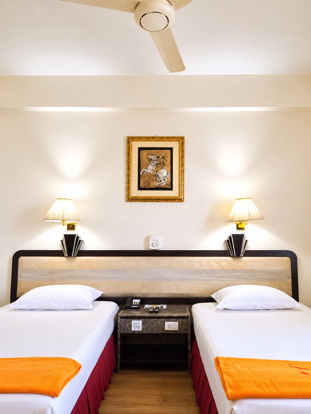 A hotel room with two beds, a ceiling fan, bedside lamps, a framed artwork on the wall, and a small table with a phone and magazines.