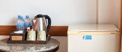 A compact refrigerator with a sticker, a small table with a tray holding water bottles, a kettle, and a mug, and a wooden chair.