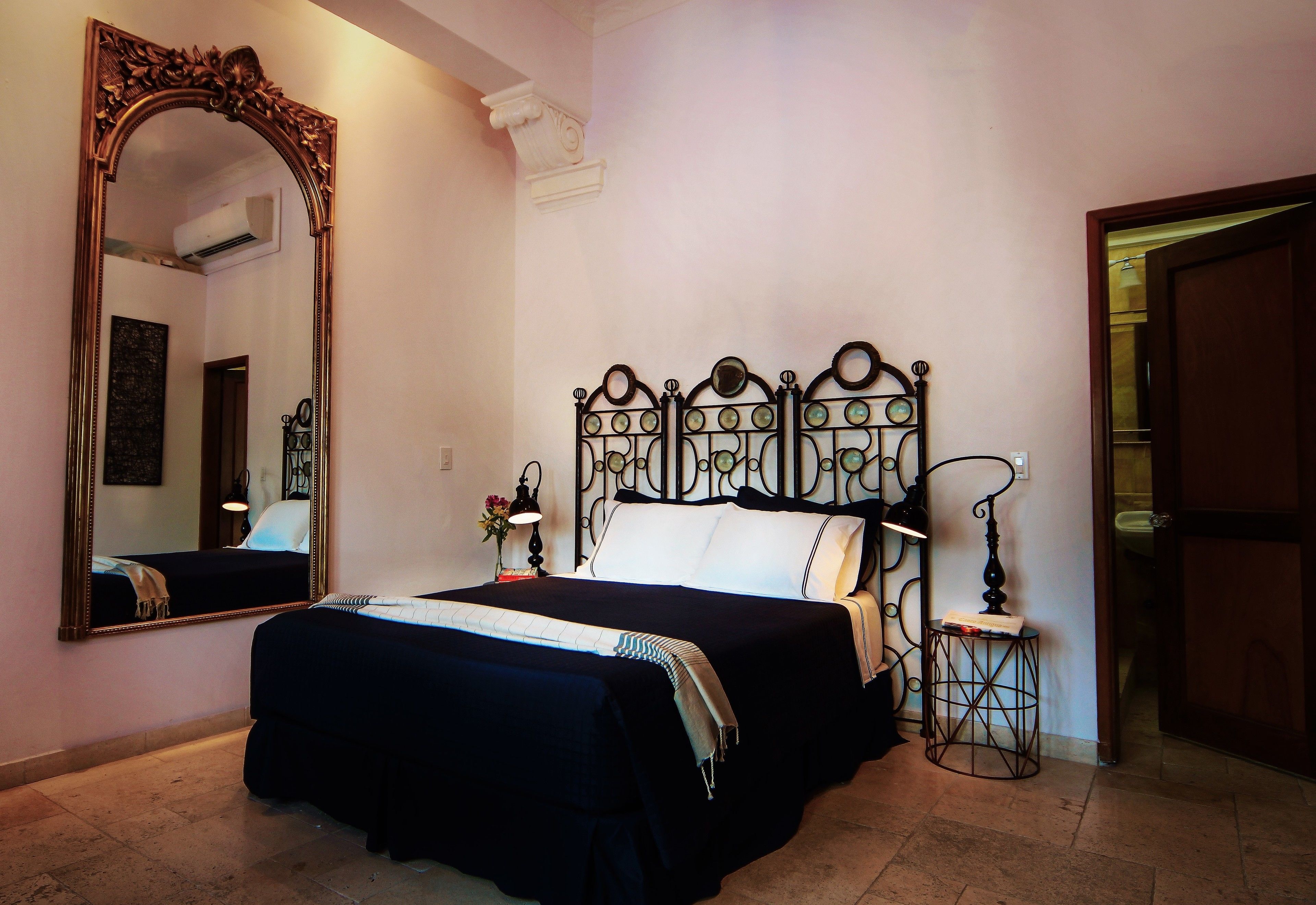 A bedroom with a large ornate mirror, a bed with a dark-colored bedspread, a bedside table with a lamp, and a door leading to another room.