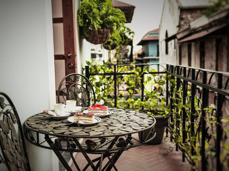 A round metal table with plates of food and cups, set on a balcony with a metal railing and potted plants.
