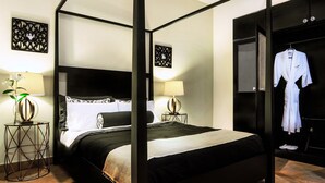A four-poster bed with a black and white bedspread, two bedside tables with lamps, and a robe hanging on a rack. - La Isabela Suites (Panama City)