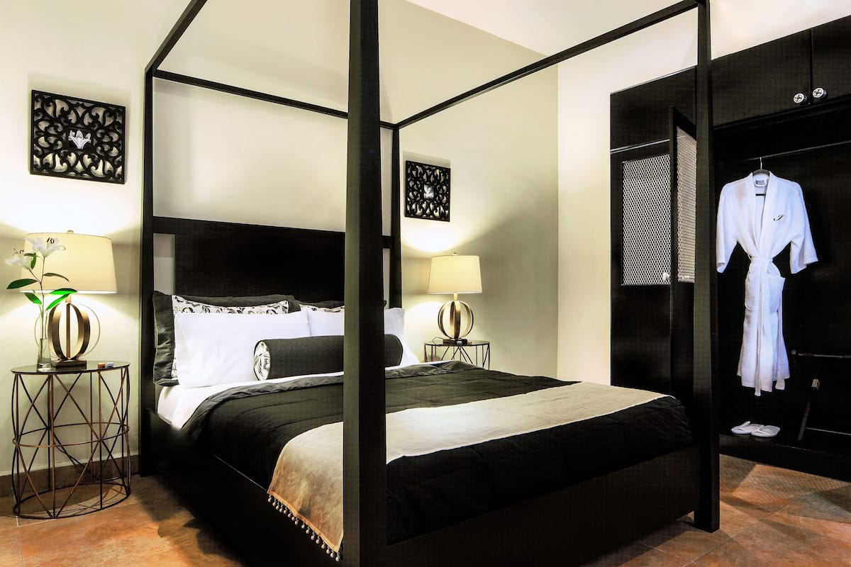 Una cama con dosel, con un edredón blanco y negro, dos mesitas de noche con lámparas, y una bata colgada en un perchero.
