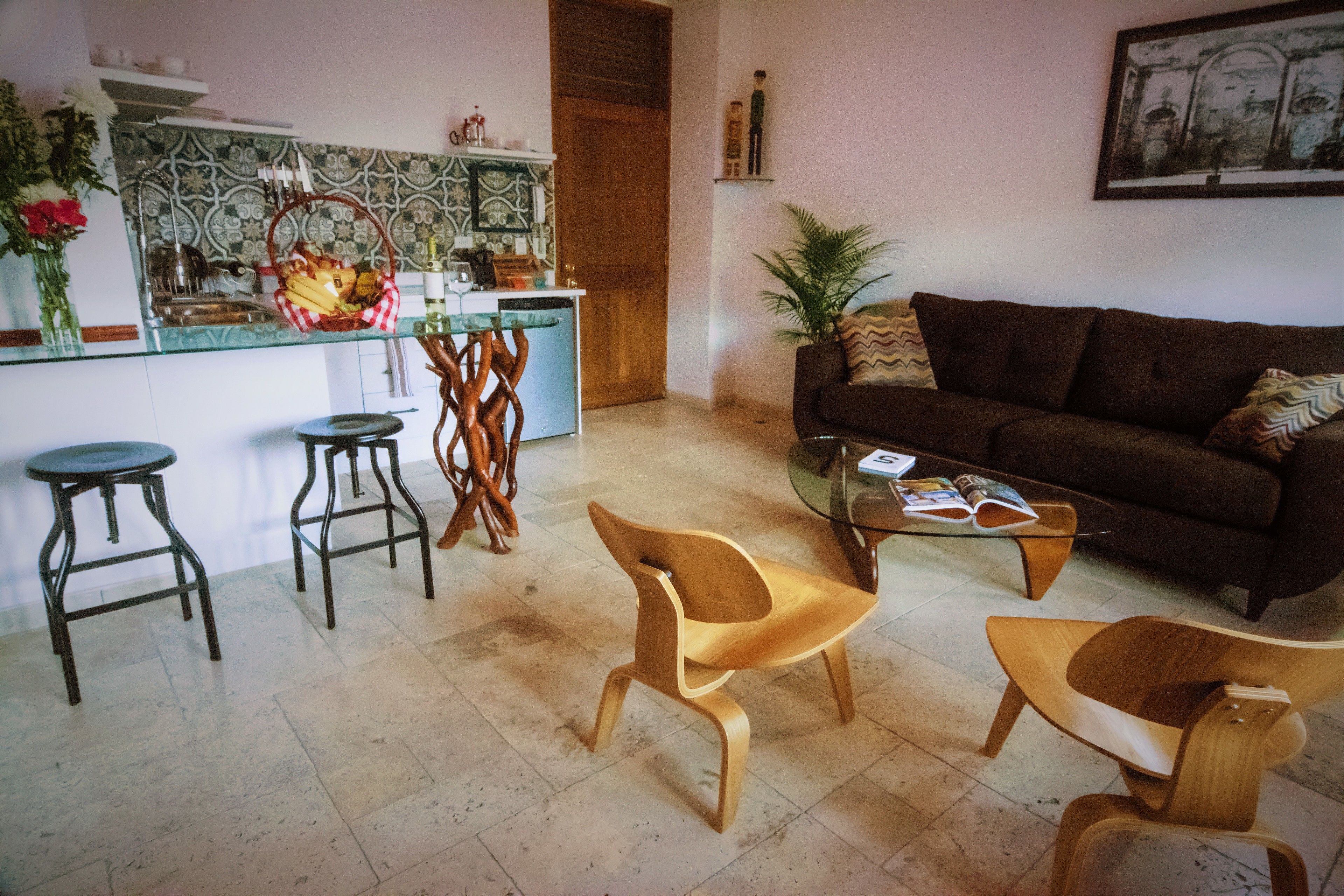 A living room with a brown sofa, two wooden chairs, a glass coffee table, and a kitchen area with a tiled backsplash and a wooden door.